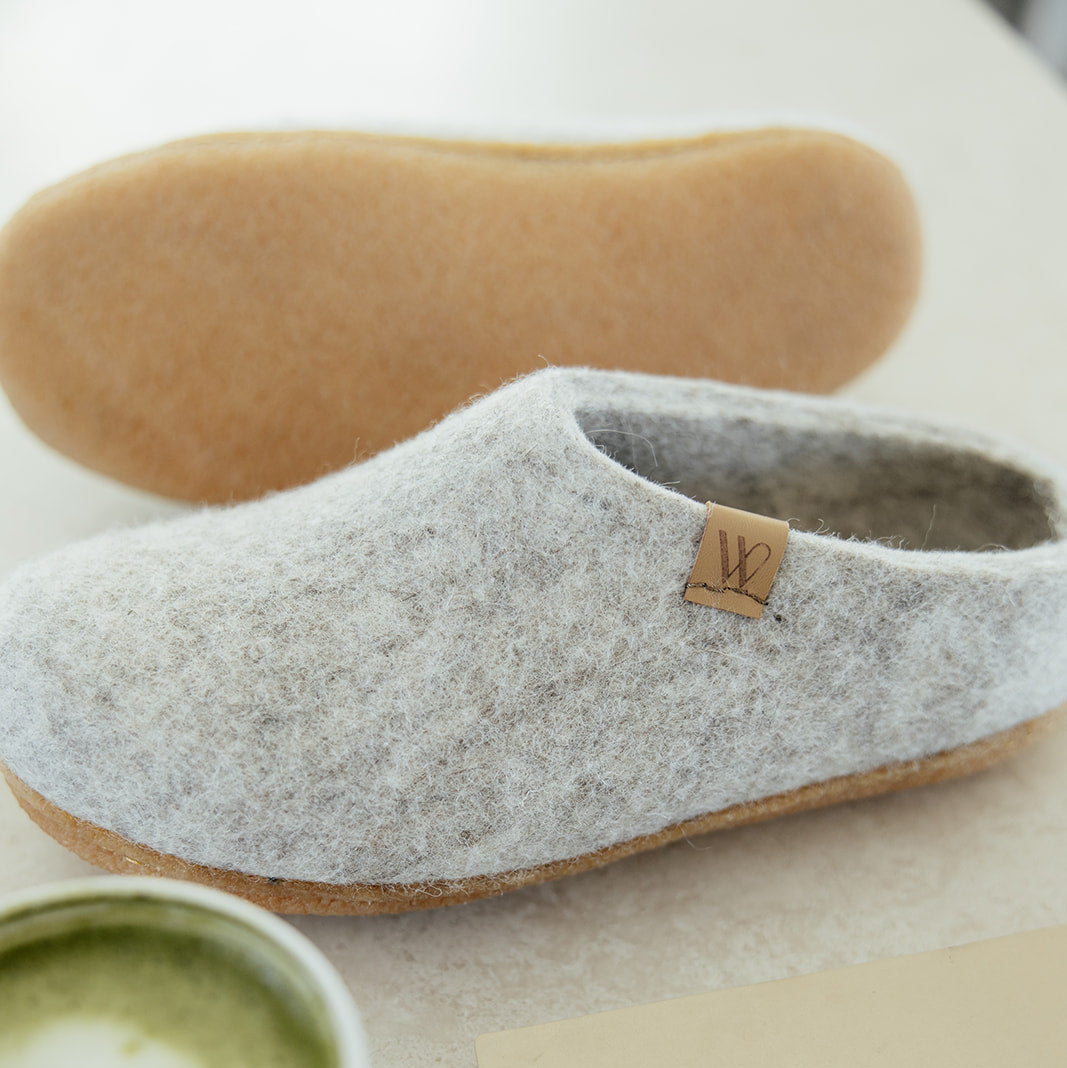A pair of light gray felt slip-on slippers with natural cork soles, styled on a light beige surface with a cup of matcha latte, a notebook, and the other slipper's sole visible in the background.