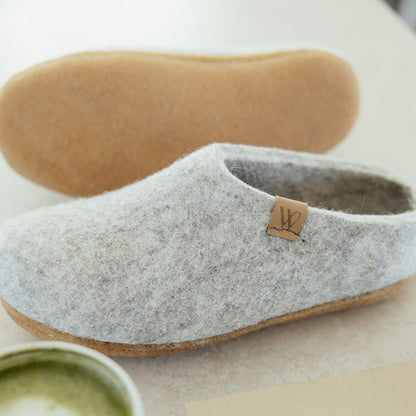 A pair of light gray felt slip-on slippers with natural cork soles, styled on a light beige surface with a cup of matcha latte, a notebook, and the other slipper's sole visible in the background.