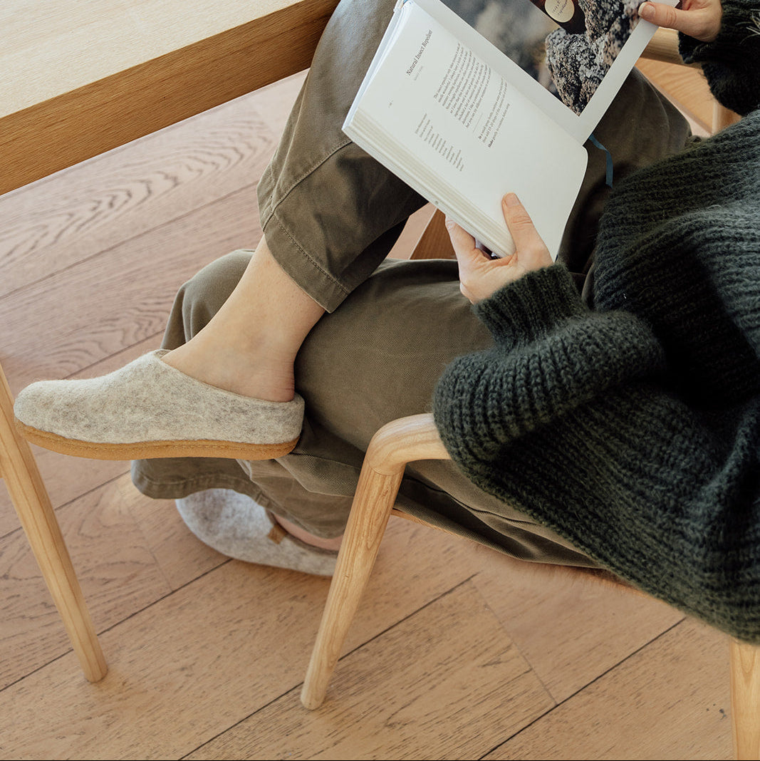A person sitting in a wooden chair on a light wood floor, holding an open magazine. The person is wearing a dark green knit sweater, olive pants, and beige slippers, with a dark green mug on a wooden ledge behind them.