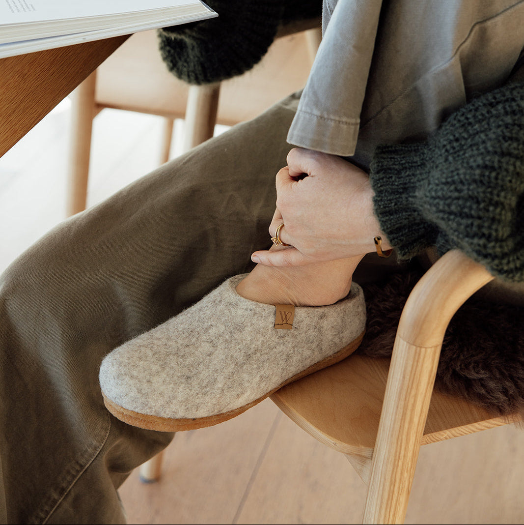 A person sitting in a wooden chair at a table, putting on a light beige felt slipper. The person is wearing a dark green chunky knit sweater and olive green pants, with an open book in one hand and a mug on the table in the background.