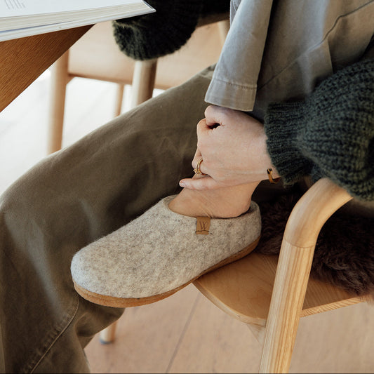 A person sitting in a wooden chair at a table, putting on a light beige felt slipper. The person is wearing a dark green chunky knit sweater and olive green pants, with an open book in one hand and a mug on the table in the background.